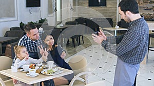 Amiable waiter take photo of friendly young family in cafe on smartphone.