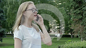 Amiable youthful woman in spectacles answering briefly on a phone call spending time outside on sunny summer day. Camera