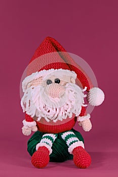 Amigurumi one Christmas gnome with red hat and white beard sits on pink background. Soft DIY toy made of natural cotton