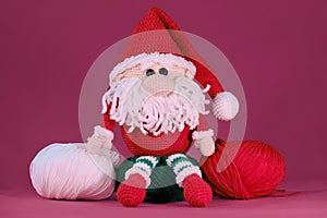 Amigurumi one Christmas gnome with red hat and white beard sits on pink background. Soft DIY toy made of natural cotton