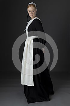 An Amish woman wearing a long black dress, apron, and cap against a studio backdrop
