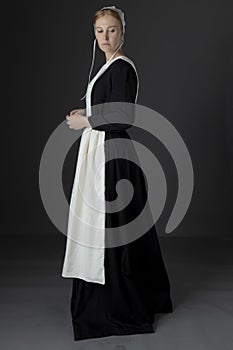 An Amish woman wearing a long black dress, apron, and cap against a studio backdrop