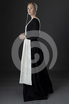 An Amish woman wearing a long black dress, apron, and cap against a studio backdrop