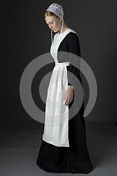 An Amish woman wearing a long black dress, apron, and cap against a studio backdrop