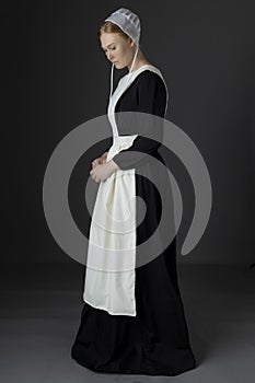 An Amish woman wearing a long black dress, apron, and cap against a studio backdrop