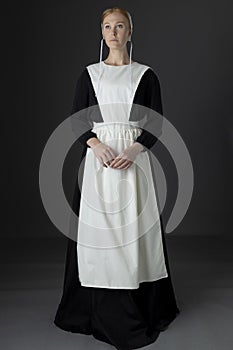 An Amish woman wearing a long black dress, apron, and cap against a studio backdrop