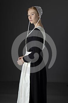 An Amish woman wearing a long black dress, apron, and cap against a studio backdrop