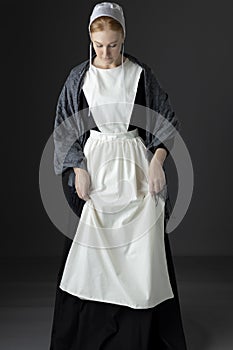 An Amish woman wearing a long black dress, shawl, apron, and cap against a studio backdrop