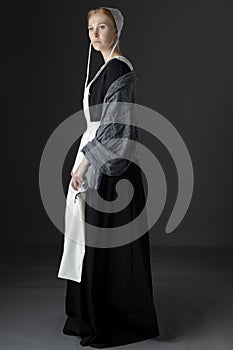 An Amish woman wearing a long black dress, shawl, apron, and cap against a studio backdrop