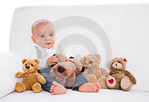 Amongst toys: cute baby sitting on white sofa with teddy bears