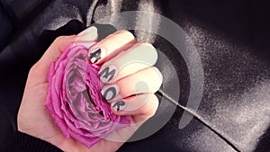 AMOR word on nails manicure hold Pink rose flower on black silk fabric. Minimal flat lay nature. Female hand. Love