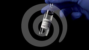 Ampoule of coronavirus vaccine on a black background in the hands of a doctor in blue medical protective gloves