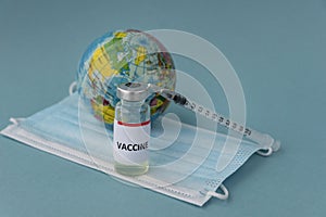 Ampoule with vaccine and inscription on it, syringe and little toy globe on medical mask on blue background, copy space.