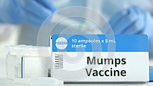 Ampoules with mumps vaccine on the table near working laboratory assistant, fictional logo on the box
