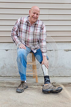 Amputee man on a stool looking forward reaching down to adjust prosthetic leg