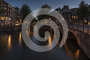 Amsterdam canal and bridge, at dusk. The canal is calm and the lights around the bridges arches are reflecting on the water