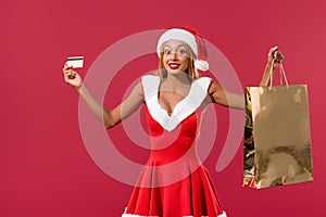 African american girl in santa hat and christmas dress holding credit card and shopping bag isolated on red