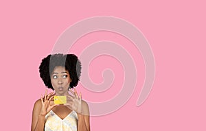 Amused African American woman with curly hair playfully holding a credit card to her lips