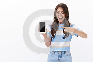 Amused cheerful young woman introduce smartphone app, hold mobile phone, pointing and looking display, smiling excited