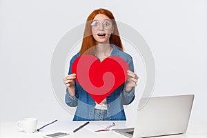 Amused and dreamy surprised redhead girl received big red valentines day heart from coworker, standing near working