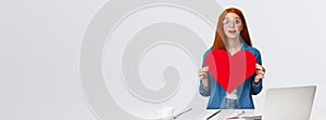 Amused and dreamy surprised redhead girl received big red valentines day heart from coworker, standing near working