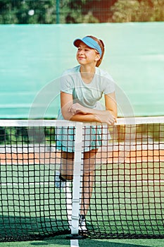 Amused happy girl leaning on a tennis court net in a light blue sportswear
