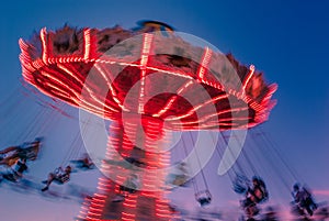 Amusement park motion blurred riders on brightly lit retro vintage tilting swing at blue hour.