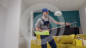 Amusing man with beard from cleaning service washing the floor in the kitchen and toying with the mop as a guitar
