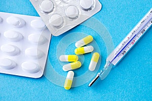 Analog thermometer on blue background with bunch of medicaments in white/yellow capsules