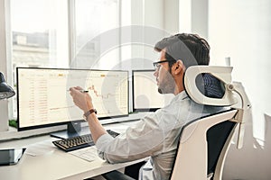Analyzing data. Back view of young businessman or trader pointing on the data on computer screen with pen while working