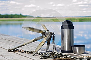 Anchor with a thermos of tea on a background of nature are at the pier