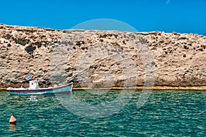 Anchored small fishing boat under Greek flag, azure sea waters, clear blue sky, rocky hill, red fishing buoy floating