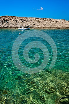 Anchored small fishing boat under Greek flag, azure sea waters, clear blue sky, rocky hill, red fishing buoy floating