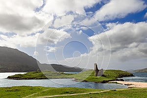 An ancient Ardvreck Castle ruin stands on a lush green islet under a dynamic sky, by the tranquil waters of Loch Assynt