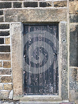 Ancient black wooden door with old faded peeling paint in a heavy stone frame with a closed bolt and metal hinges