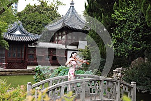 Ancient historic Asian Chinese woman artist traditional chi-pao cheongsam in a garden play musical instruments violin on bridge