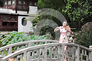 Ancient historic Asian Chinese woman artist traditional chi-pao cheongsam in a garden play musical instruments violin on bridge