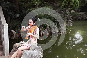 Ancient historic Asian Chinese woman artist traditional chi-pao cheongsam in a garden play musical instruments violin on bridge