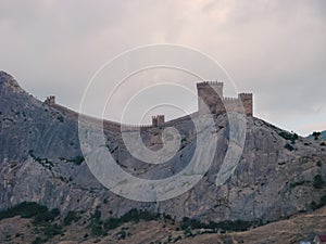 An ancient medieval fortress on the edge of a cliff is surrounded by large, bright clouds.