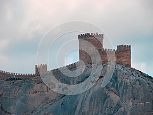 An ancient medieval fortress on the edge of a cliff is surrounded by large, bright clouds.