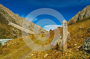 Ancient milepost beside a trail through a vivid autumn meadow, beneath towering mountains, expansive blue sky.