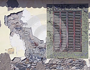 an ancient red wooden shuttered window in a frame with peeling faded green paint in an old house wall with broken cracked cement
