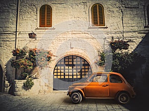 Ancient small orange Italian car, parked on the road in front of an ancient dwelling.