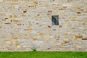 An ancient stone wall of hewn stone with a small window. Green grass