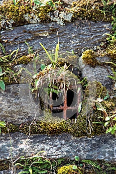 Ancient wall made of natural rocks with window-like water-drain pipe in the center and with plants and lichens between blocks