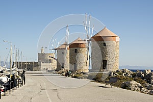 Ancient windmills on stony Rhodes coastline in harbor, old historic buildings, place of interest, blue sky