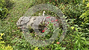 An ancient woman sculpture over grass with moss and red flowers at Colombian San Agustin archaeological park.