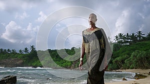 Androgynous black person in luxury dress walks along scenic ocean beach slowmo. Gender fluid ethnic fashion model