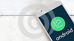 Android 12 logo on a screen of a mobile on a white wooden table.