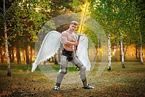 Angel, a bodybuilder in plate armor on his legs with wings behind his back, holds in his hands a fiery two-handed sword